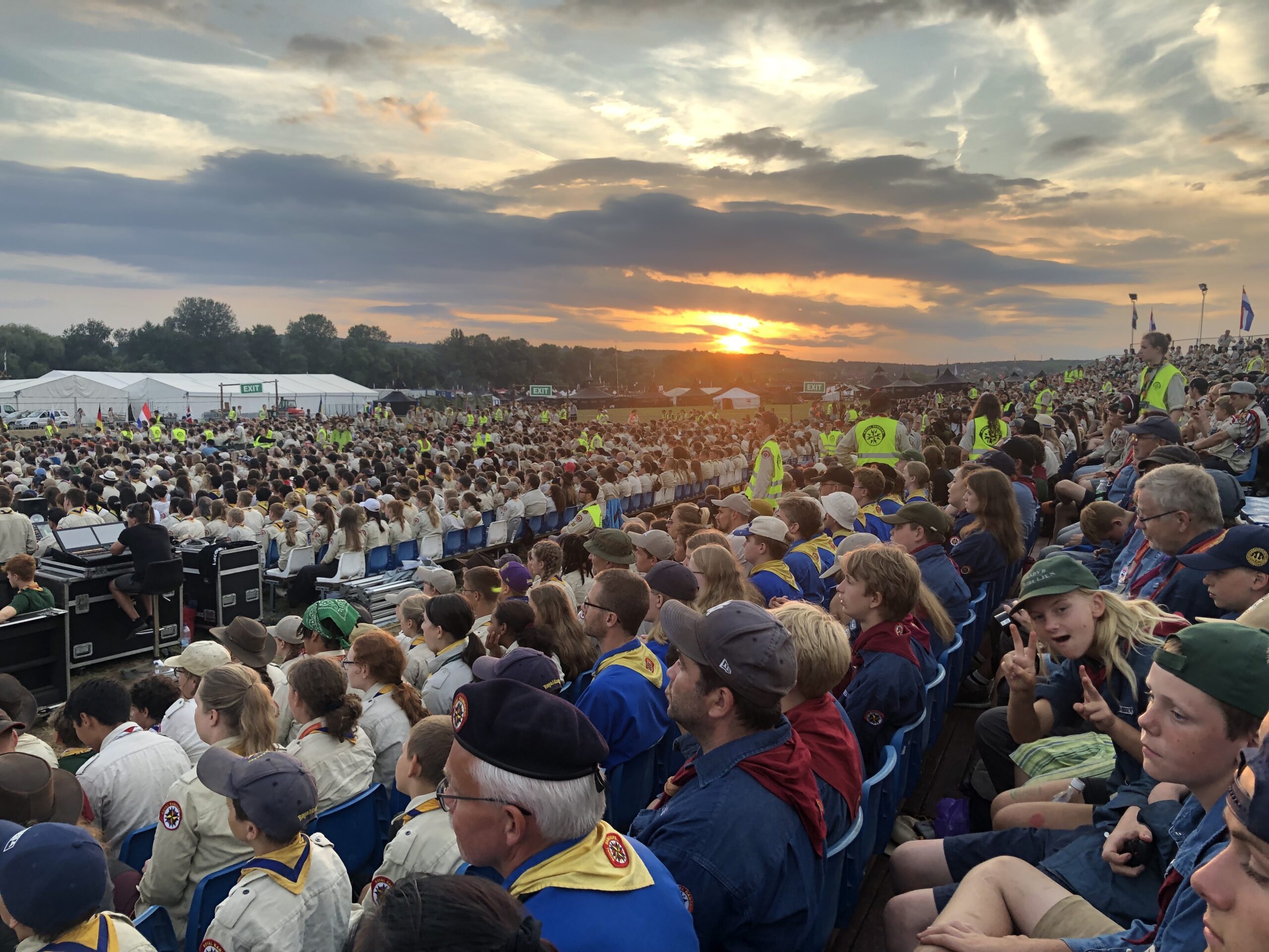 Mange hundre speidere sitter på et stadion, solnedgang i horisonten bak skyer